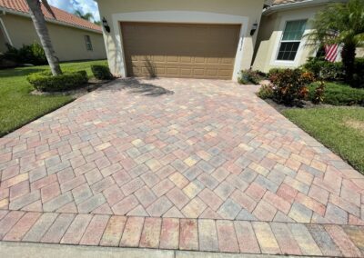 A house with a tan garage door and a neatly paved driveway made of interlocking brick in a herringbone pattern. The front yard has well-manicured grass, a palm tree, and various shrubs and flowers. An American flag is visible near the side of the house.