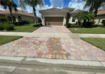 A house with a light brown tiled roof and beige exterior has a driveway made of multicolored brick pavers. Palm trees and manicured lawn surround the house. A blue sky with scattered clouds is visible in the background.