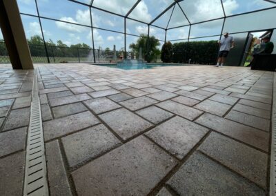 A spacious outdoor patio with tan tiles, featuring a small pool and jacuzzi in the center, surrounded by a screened enclosure. Two people stand near the edge of the patio under a mostly cloudy sky, with greenery visible outside the enclosure.