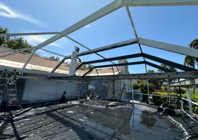 A worker in protective clothing and mask stands on a ladder, spray painting the framework of a large outdoor structure. The ground is covered with black plastic sheeting, and the surrounding area features a house with a tiled roof and green foliage.