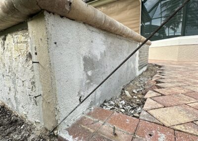 A close-up of a house foundation reveals a damaged stucco facade with cracks, missing sections, and exposed concrete. An iron rod supports the structure, and nearby pavers on a brick patio are misaligned and partially removed, exposing the dirt beneath.