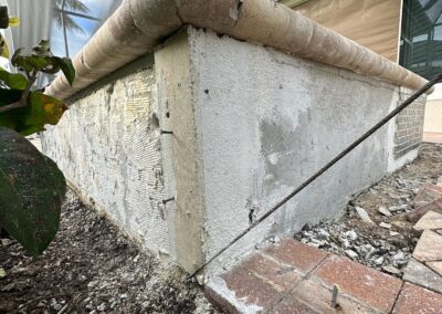 View of a partially damaged stucco wall corner with exposed reinforcement wire and deteriorated concrete. The base of the wall has chipped stucco and surrounding soil appears disturbed. Bricks are seen on the ground beside the wall.