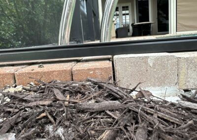 Close-up of a patio edge showing a transition between mulch and paving stones. The paving stones form a small wall separating the garden bed with mulch from the patio area. In the background, a glass door and patio furniture are visible.