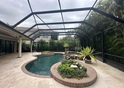 A screened-in patio features a small, curvy swimming pool with a central plant island. Adjacent to the pool is a hot tub with stone detailing and a waterfall feature. Lush greenery surrounds the patio, including large plants and trees. The sky is overcast.