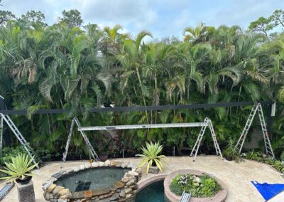 A backyard with an in-ground pool and a circular hot tub surrounded by stone. There are lush green palm trees in the background and several ladders and construction equipment set up, indicating ongoing work. The patio is paved with stone tiles.