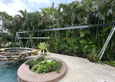 A partly cloudy sky overlooks a backyard with a pool and hot tub. In the foreground, there's a brick planter with lush greenery. Two ladders and a long horizontal beam suggest ongoing construction or maintenance near tall tropical plants along the fence.