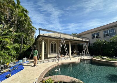 Construction workers build a patio cover beside a swimming pool in a backyard. Ladders are positioned under a steel frame being erected, while tools and materials are scattered around. Tall palm trees and a two-story house are in the background on a sunny day.