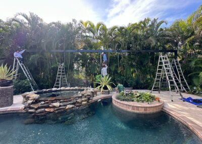 Four people are installing a structure near a pool surrounded by lush greenery. Three ladders are set up, with one person on each ladder. The fourth person is standing on a rocky platform next to a small waterfall feature in the pool.