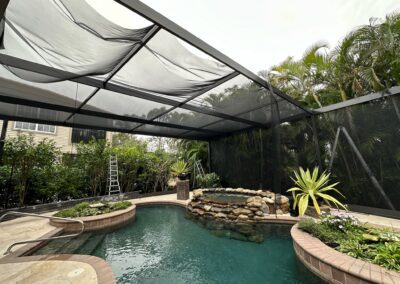 A backyard features a screened-in pool area with a hot tub, a stone waterfall, and various potted plants. A ladder is propped up against the screen enclosure, with lush greenery and trees visible beyond the screened area.