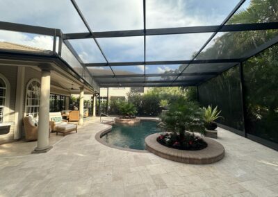 A luxurious screened-in patio featuring a kidney-shaped pool surrounded by potted plants and tropical greenery. There are cushioned wicker chairs and a sofa under a covered area supported by columns. The ceiling consists of a metal frame with mesh screening.