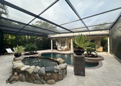 A modern outdoor patio featuring a pool surrounded by a stone border with large potted plants. The area is enclosed by a screened structure. Lounge chairs and seating areas with cushions are arranged around the patio, framed by lush greenery in the background.