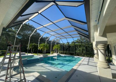 An outdoor pool area enclosed by a black screened structure. The pool features both shallow and deep sections, with a hot tub and water features. A metal ladder is positioned on the left side. Trees and greenery are visible through the screens.