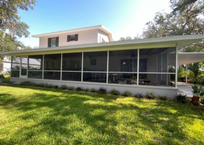 A two-story house with a screened-in porch stretching along the back. The porch has a fan and furniture within it, and the surrounding yard is green with grass, plants, and trees. The bright, clear sky highlights a serene outdoor environment.