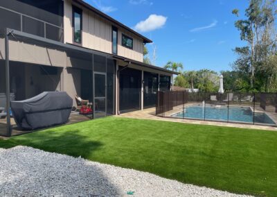 A backyard with a large rectangular pool enclosed by a black safety fence. Adjacent to the pool is a screened patio area with outdoor furniture and a covered grill. The yard features a green lawn and a gravel pathway, with a modern two-story house in the background.