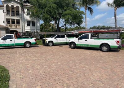 Three white pickup trucks with green trim and "Superior Roof" branding are parked on a brick driveway in front of a large white house with arched windows and palm trees in the background. The trucks have ladders mounted on top.